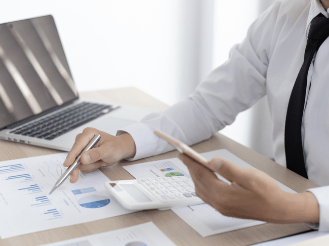 4 A business professional in a white shirt and black tie analyzing financial charts and graphs at a desk, holding a smartphone in one hand and a pen in the other. A calculator and an open laptop 