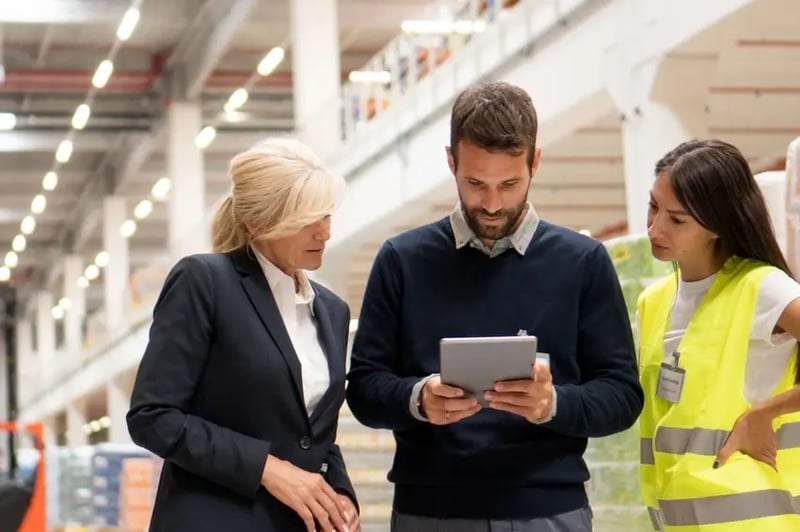 Operations_Leaders Three professionals in a warehouse discussing logistics while reviewing information on a tablet. A man in business attire holds the tablet, while a woman in a suit and another in a high-visibility vest listen attentively.